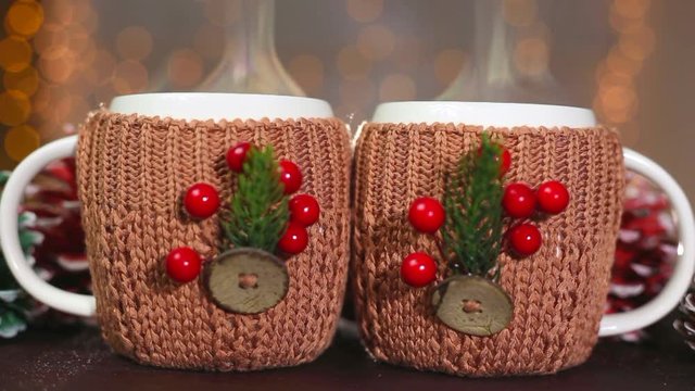 Closeup of two white steaming winter cups in handmade brown cute knitted covers with festive christmas decorations at blurry lights of orange garland background. Real time full hd video footage.