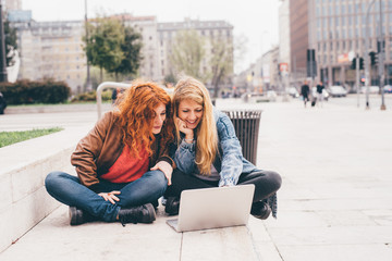 Two young beautiful blonde and redhead caucasian women outdoor sitting using computer – small business, technology, sharing concept