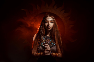 Beautiful, red-haired girl - a priestess of the sun. Dress and jewelry in ethnic style. Unusual make-up in orange and gold tones. Girl praying in silence.  Fantastic photography