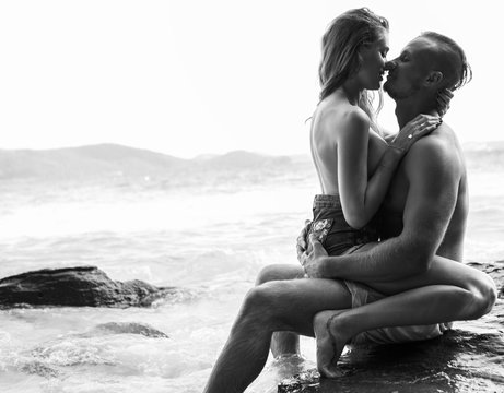 Attractive young topless couple hugging while sitting on the rock by the sea over sky background - black and white photo