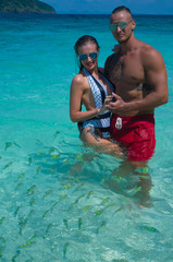 Attractive young couple wearing swimsuits and sunglasses looking at the camera and smiling while standing in the lagoon sea with colorful fishes over beautiful blue sky background