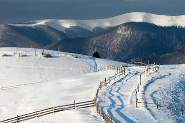 Pasture fence on snowy hills in the sunlight against the background of a mountain range and a dark sky. A fabulous winter landscape.