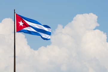 Cuban flag flying from a flagpole in the wind in front of a soft blue sky with tropical clouds in Havana, Cuba