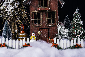 miniature Christmas scene on rustic house and decorated white picket fence in deep snow with snowman and pine trees