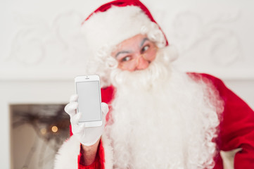Obraz na płótnie Canvas Santa Claus shows a smartphone against fireplace