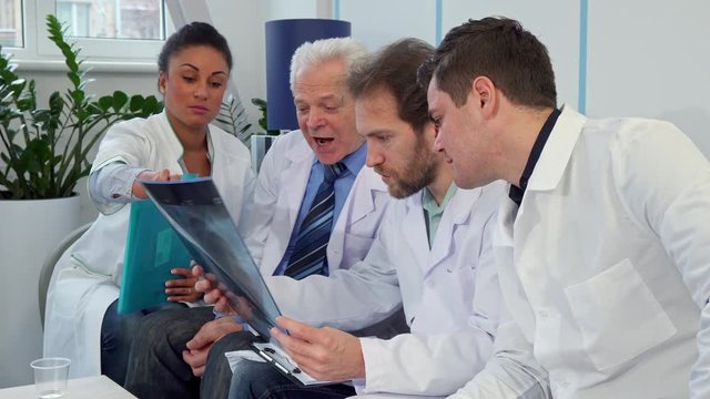 Medical team discussing x-ray image of human's rib cage. Bearded middle aged man in white coat holding x-ray in his hand. Three caucasian male doctors and one african american female doctor sitting on