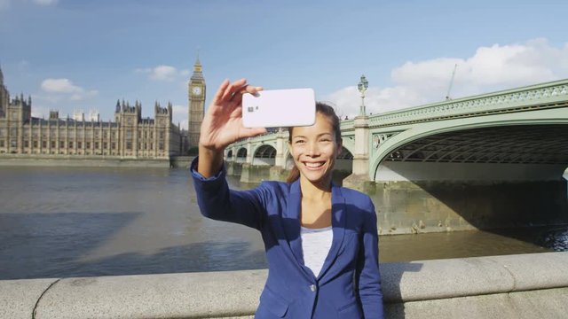 Business woman taking selfie using phone in London on business travel. Businesswoman smiling at camera with Thames river, Big Ben and Westminster bridge background. Asian woman using smartphone.