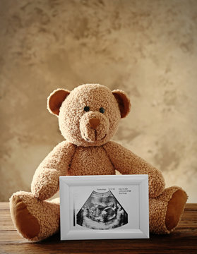 Teddy bear holding photo frame with ultrasound picture of baby on table