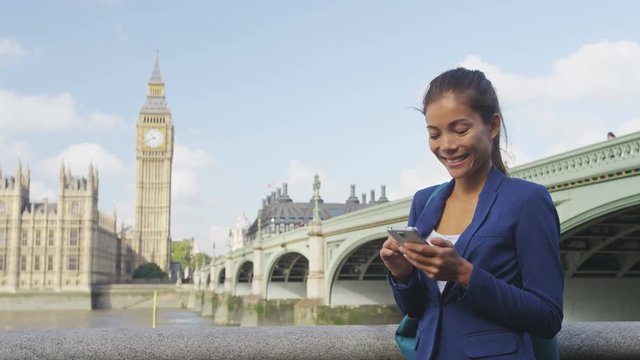 Businesswoman using app on smart phone by Westminster Bridge, London, England. Young business woman using smartphone smiling happy wearing suit jacket outdoors. Urban female professional, 20s.