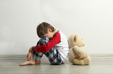 Sad little boy with teddy bear sitting on floor in empty room