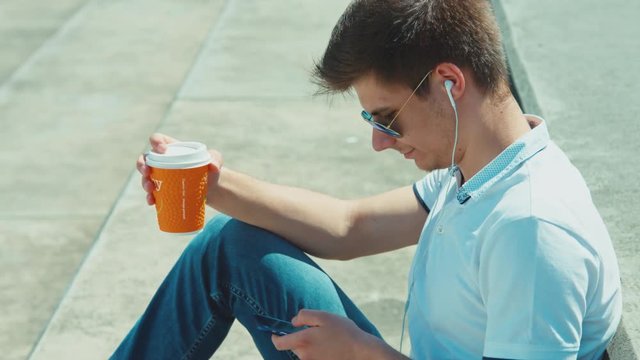Closeup portrait student male in sunglasses using cellphone and holding cup of tea sitting on outdoor in urban park