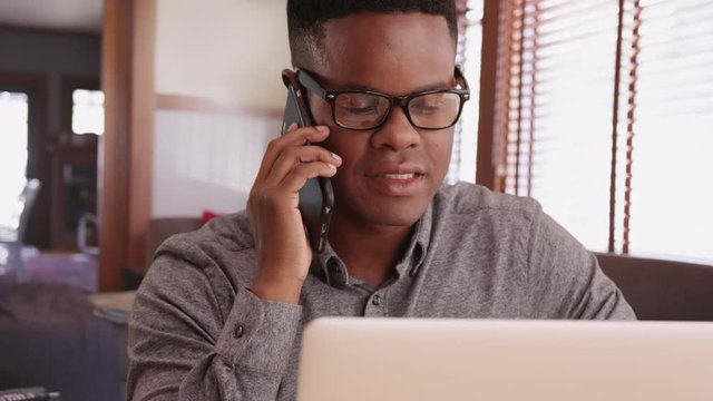 Black male with glasses talks on the phone working on his laptop