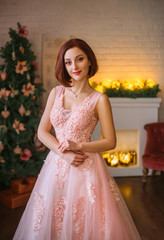 Fototapeta premium Seductive girl in the lush, pink dress standing in the room. Christmas atmosphere fills the picture. Gentle make-up, simple hairstyle.