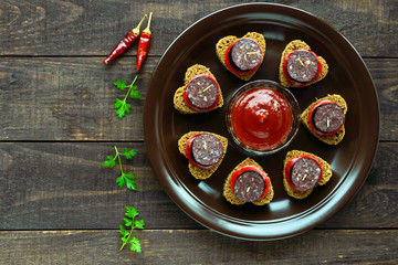 Sandwiches with black rye bread in the shape of a heart, blood sausage (Morcillo) and pieces of sweet pepper on skewers in a ceramic bowl with tomato sauce on a dark wooden background. The top view.