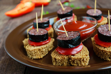 Sandwiches with black rye bread in the shape of a heart, blood sausage (Morcillo) and pieces of sweet pepper on skewers in a ceramic bowl with tomato sauce on a dark wooden background.  Close up