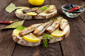 Sandwich with smoked fish capelin (sprats), lemon on black rye bread. Snack on a dark wooden table.