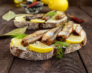 Sandwich with smoked fish capelin (sprats), lemon on black rye bread. Snack on a dark wooden table.