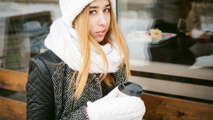 woman in white scarf and hat drinks coffee. cute young blonde girl sitting on a bench near the coffee shop, drinking takeaway coffee, holding Christmas paper cup in his hands. looking at the camera