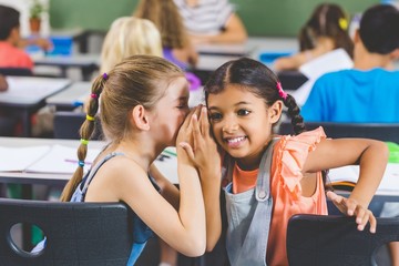 Schoolgirl whispering into her friend s ear in classroom - Powered by Adobe