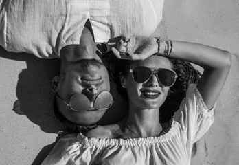 Top view closeup face of sensual pretty couple wearing white clothes and sunglasses smiling while laying on the sandy beach - black and white photo