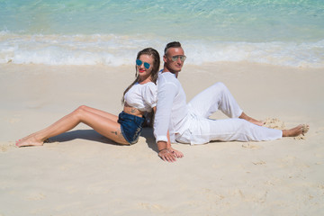 Happy young couple wearing white clothes and sunglasses smiling and looking at the camera while sitting on the sandy beach over lagoon sea and blue sky background