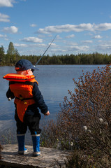 Young boy is fishing wiht his rod wearing life jacket by the lake