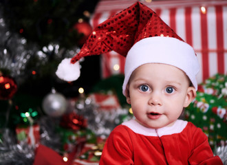 9 month old baby boy looking at the camera while wearing a santa claus outfit, with christmas presents and gifts behind him under the decorated christmas tree.