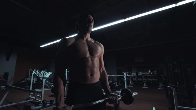Muscular man doing crossfit training in a dark shadowy gym lifting weights holding a barbell at waist level in a health and fitness concept