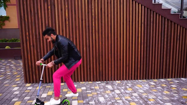 Prepossessing Arabian Masculine Yound Guy Rides Scooter, Fun and Smiles on Background of Stairs in Hotels in Open Air.