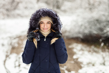 Young smiling woman wearing blue hooded real fur trim down coat enjoying view in winter forest outdoors. Nature cold season freshness concept.