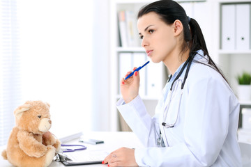 Young brunette female doctor sitting  with clipboard near window in  hospital and filling up medical history form.