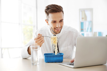 Young man eating instant noodles while working with laptop in office