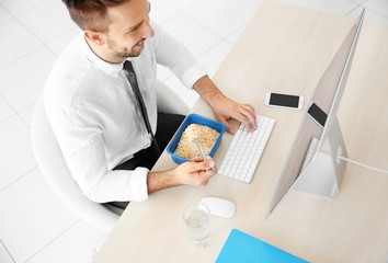 Young man eating instant noodles while working with computer in office