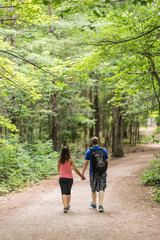 Couple of people hiking together in the woods down a dirt path on a beautiful summer day. Green canopy of trees above them. View from the back of people - full body.