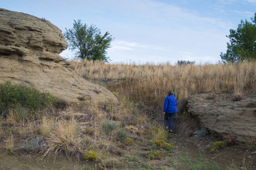 Woman in cornflower blue fleece jacket with her back to the camera climbing a path between sandstone rock formations and grassland. Thin, wispy cirrus clouds in blue sky are above.