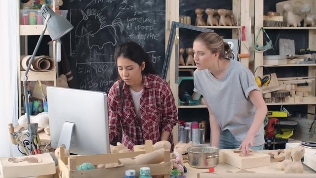 Young women standing in creative workshop looking at computer screen discussing new project and making notes