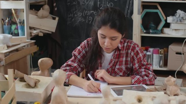 Young woman sitting in creative workshop looking at tablet and making notes about new project