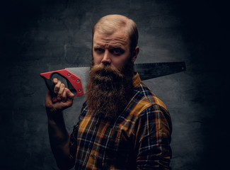 A bearded carpenter man dressed in a plaid shirt holds handsaw.