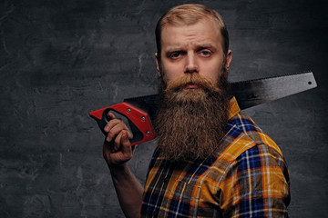 Bearded carpenter dressed in a plaid shirt holds handsaw.
