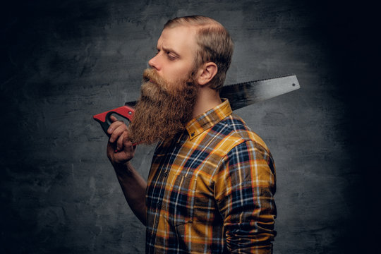 Bearded carpenter dressed in a plaid shirt holds handsaw.