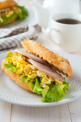 Sandwich stuffed with scrambled eggs, ham and lettuce leaves on a light wooden background. Selective focus.