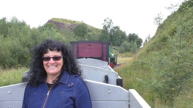 Dark curly hair woman wearing glasses shows thumbs up sign enjoying steam engine train open air wagon bumpy ride in mining area in Lake District National Park of England, United Kingdom