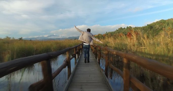 Boy with a black jacket and blue jeans is running with the hand opened like a little plane on the wooden bridge on the pond.