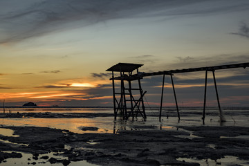 Fototapeta na wymiar Golden Hour moment,beautiful tropical sunset background, wooden water pump tower on the muddy beach. cloudy and yellow sky.surface level shot.low tide sea view