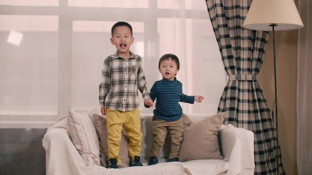 two little boy of Asian appearance, having fun on the sofa in the room, jump and laugh