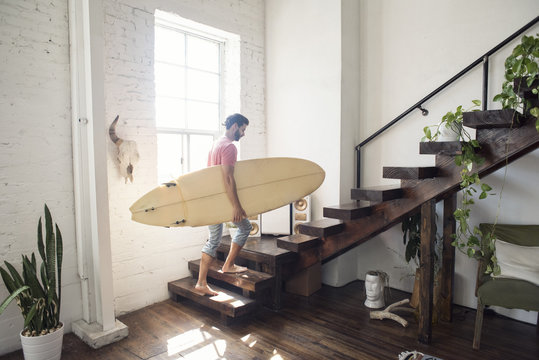 Young man carrying a surfboard on stairs in a loft