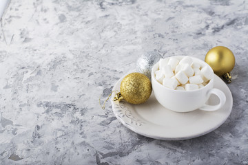 Ceramic white cup full of small marshmallow for making winter praznichnogo hot chocolate on gray concrete background surrounded by a gold Christmas ornaments balls.