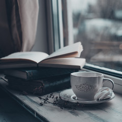 Cozy winter still life: cup of hot coffee and opened book on vintage windowsill against snow landscape from outside