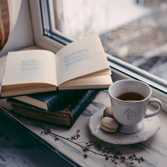 Cozy winter still life: cup of hot coffee and opened book on vintage windowsill against snow landscape from outside