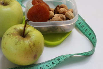 Useful food, healthy snack, lets lose weight, different nuts (cashews, walnuts) and dried apricots in a beautiful light green lunchbox to cover garden and apple on a white surface, centimeter